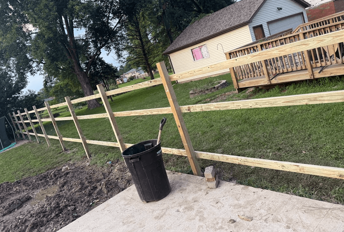 New wooden post and rail fence in a grassy backyard with a black trash can.