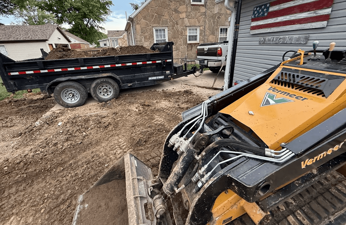 Yellow Vermeer skid steer on muddy ground next to a black trailer filled with dirt.