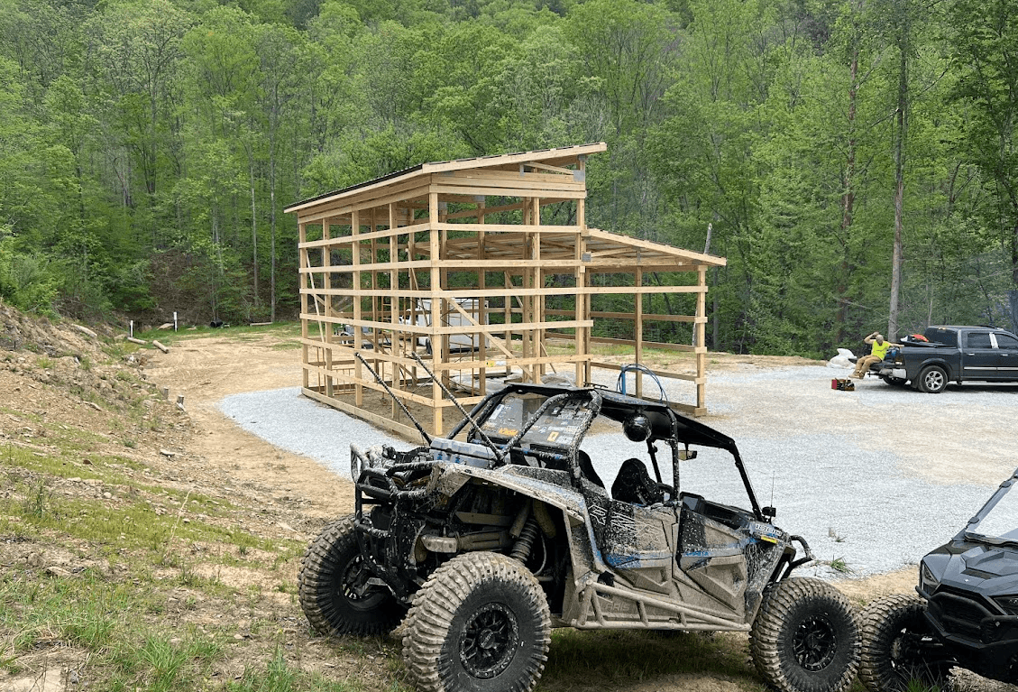 Muddy off-road vehicle parked near a wooden shed frame under construction in a forest.